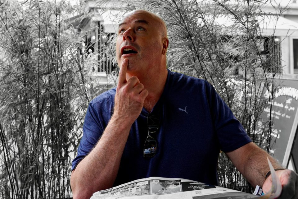Gareth at a café table with a newspaper, finger on his chin, looking up in thought. He is in colour against a black and white background.