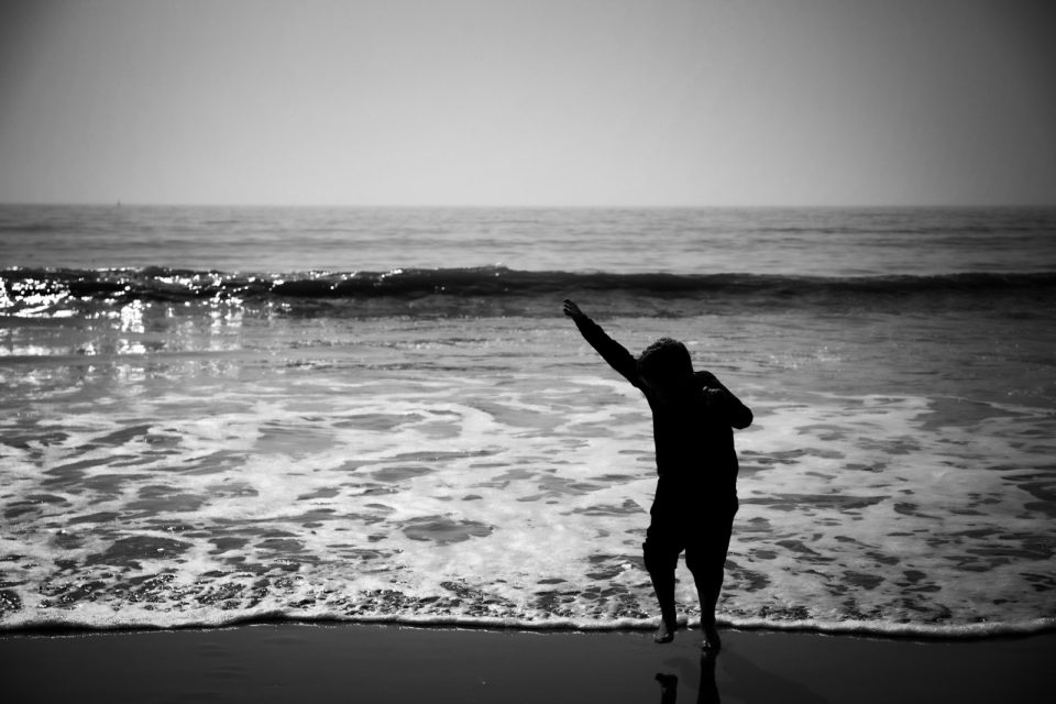 Black and white: a child silhouetted against the sea, striking a dab at the water's edge.