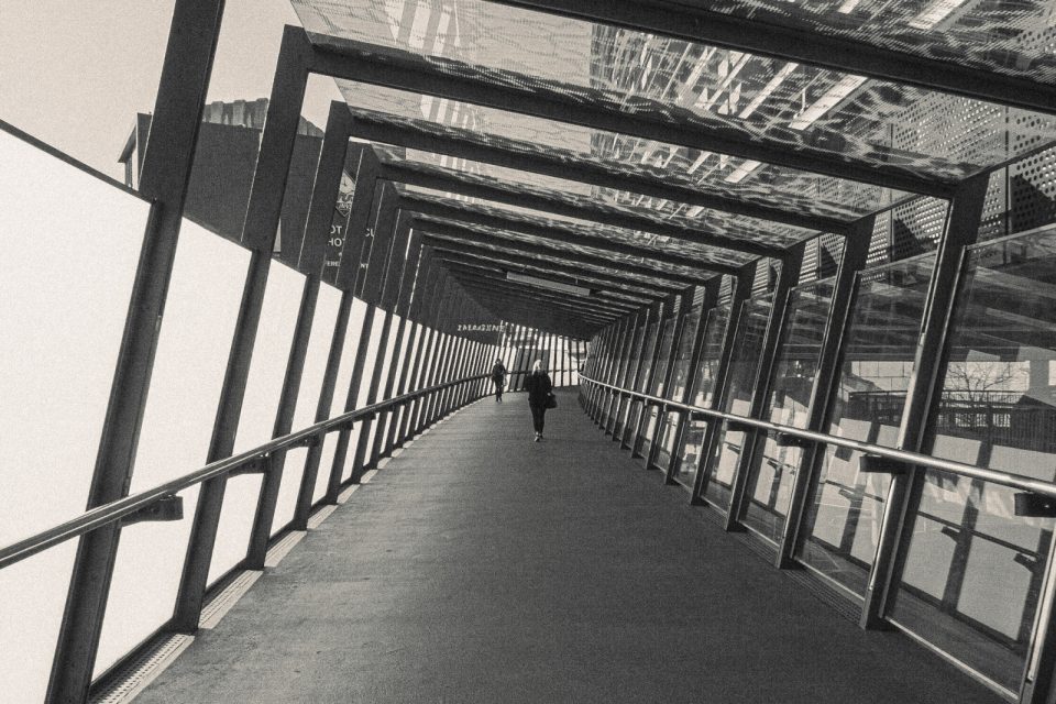 Black and white: looking down a long covered footbridge with angled steel frames, two people walking away.
