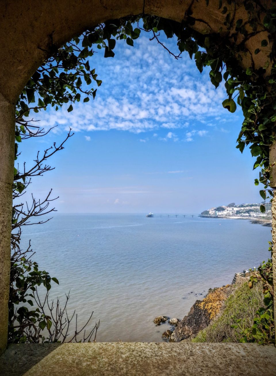 The sea and a distant pier framed by a stone arch hung with ivy, under a sky full of small clouds.