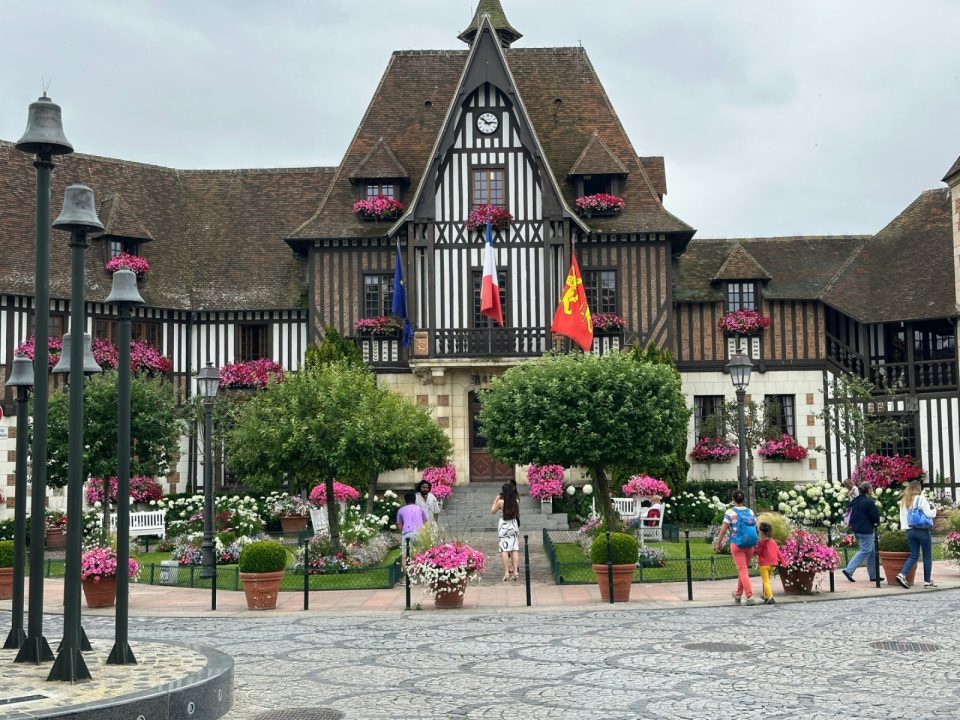 A half-timbered town hall flying the French and Normandy flags, its square full of pink flowers.