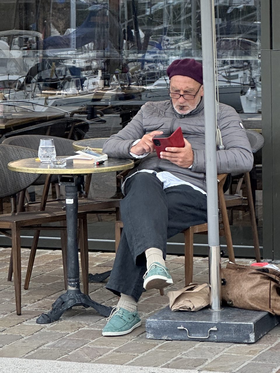 An older man in a burgundy beret reads his phone at a café table, boats reflected in the window behind him.